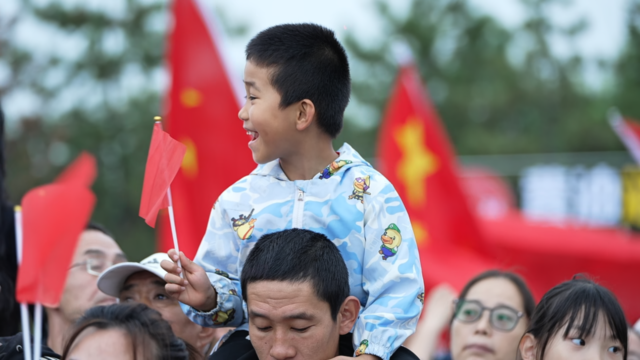 在百狮园南川古渡水路巡游表演区,一名孩童坐在家长肩头,手握小红旗,兴高采烈地观赏展演。贾玉龙摄