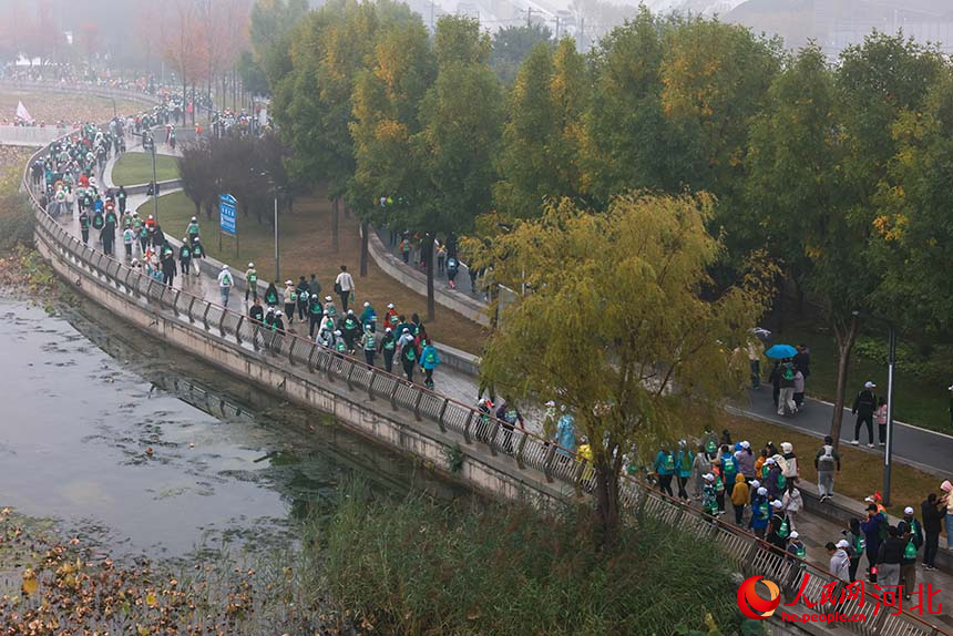 秋日细雨洒落河畔，徒步者热情不减，绵长队伍与秋日水岸风光相映成景。人民网 席彪摄